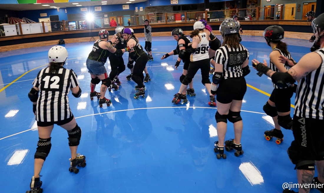 Four roller derby referees officiate from the inside of a blue track in a roller rink while gameplay happens